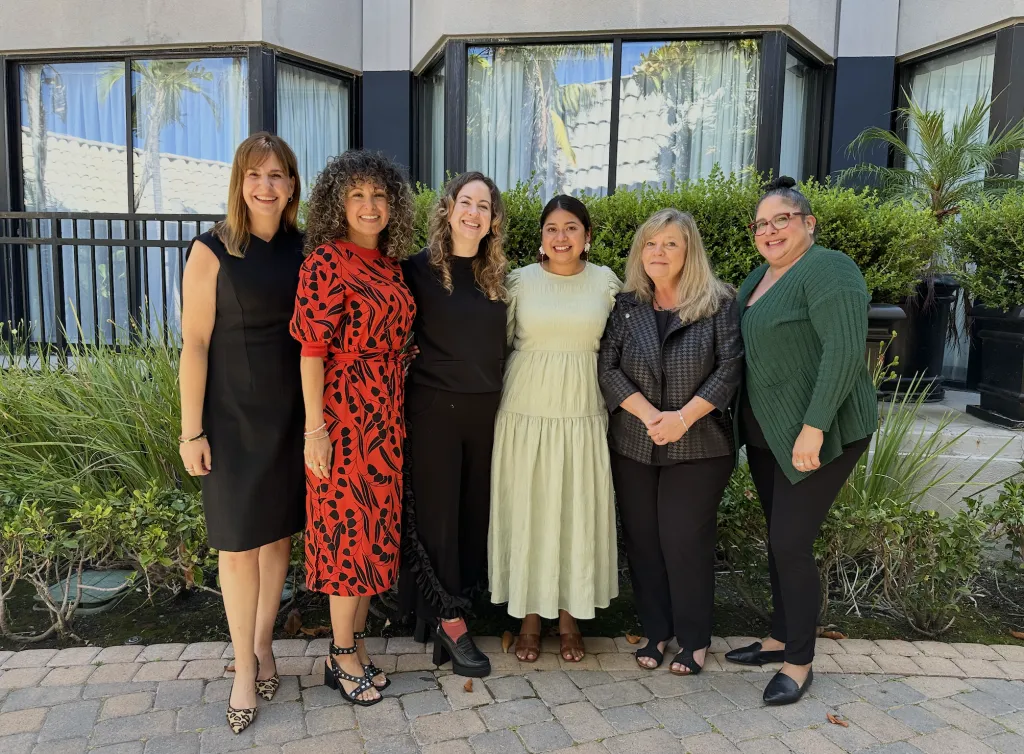 Six women are standing side by side outdoors, smiling at the camera. They are dressed in business or semi-formal attire, with greenery and a building with large windows in the background.