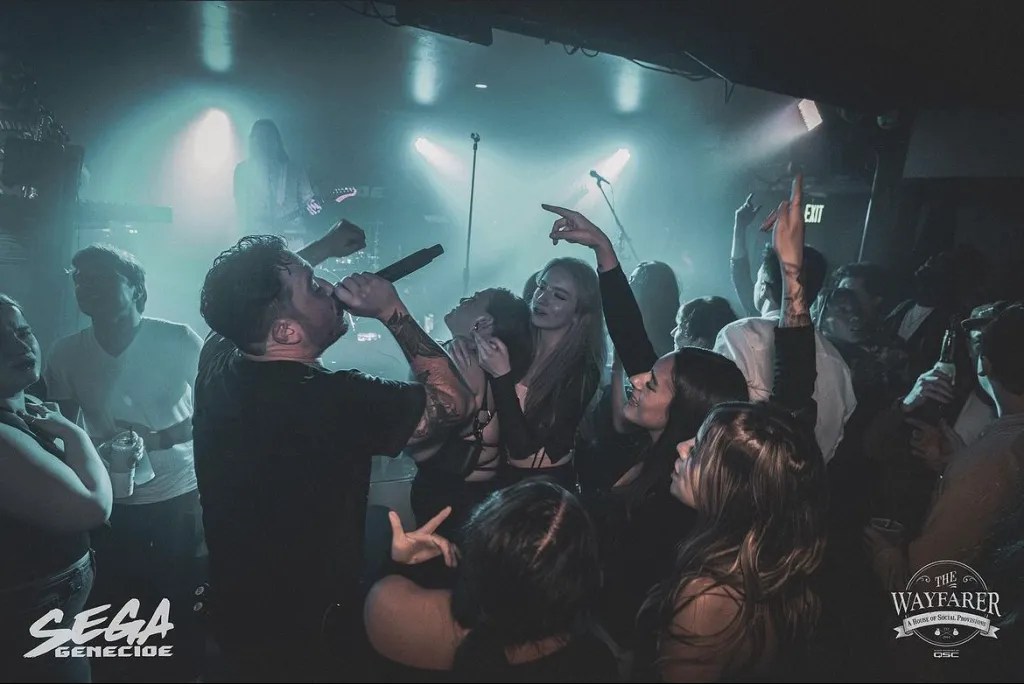 A singer performs passionately with a microphone on a crowded stage, surrounded by enthusiastic fans raising their hands in a dimly lit venue. Logos for SEGA Genecide and The Wayfarer are visible in the corners.