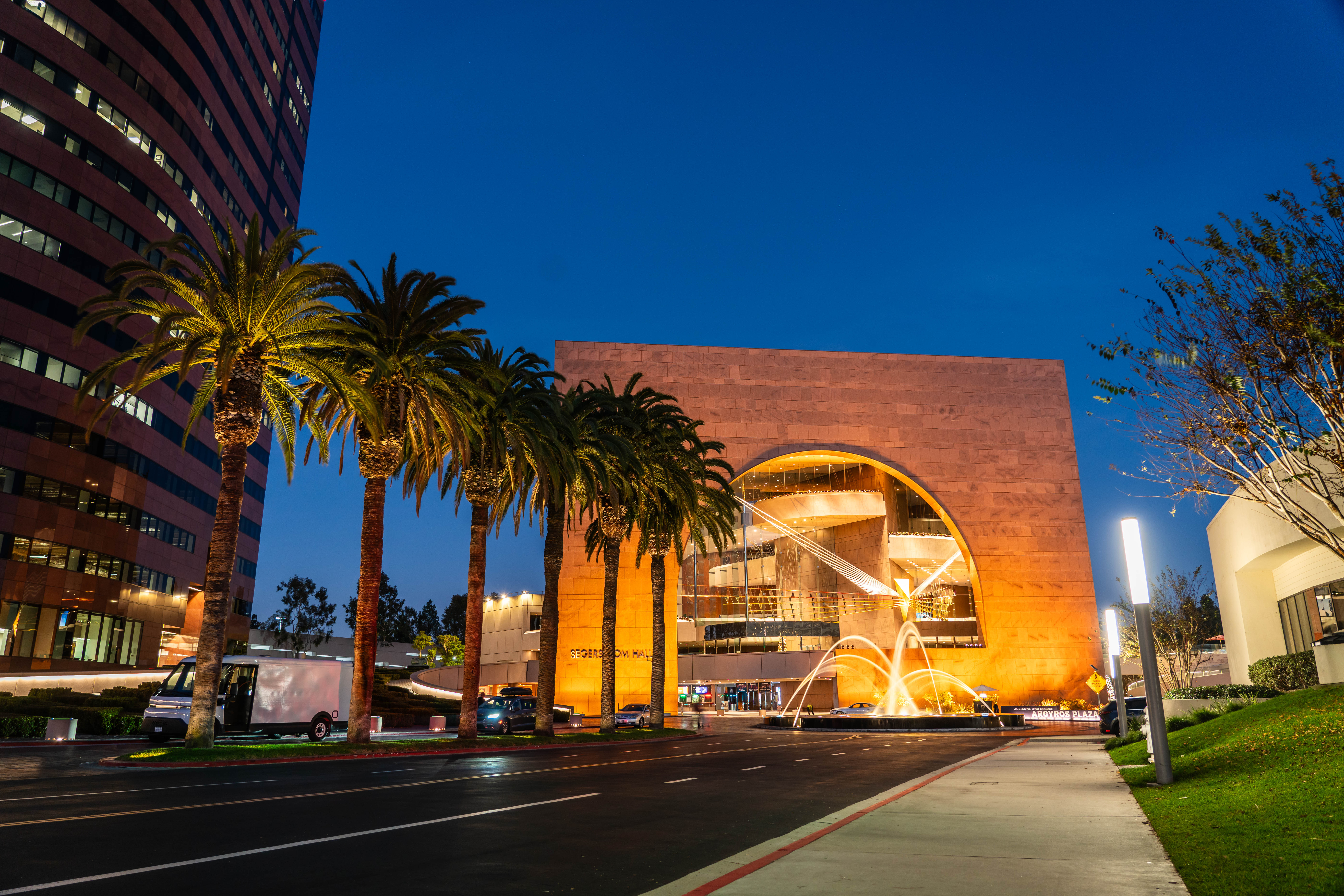 A modern building, Segerstrom Hall in Orange County, features a large circular cutout and illuminated fountain in front, surrounded by palm trees and city lights, photographed at dusk.