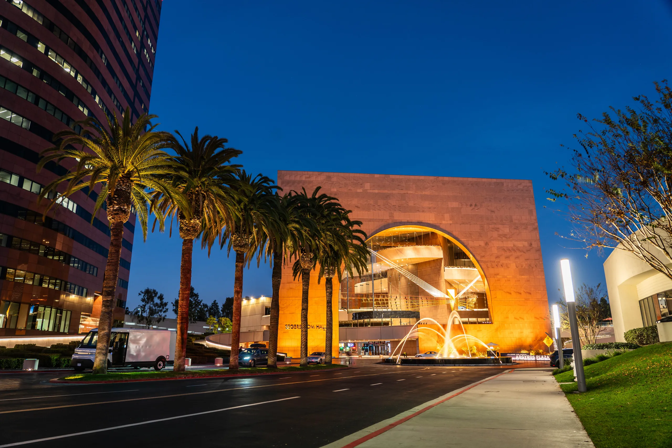 A modern building, Segerstrom Hall in Orange County, features a large circular cutout and illuminated fountain in front, surrounded by palm trees and city lights, photographed at dusk.
