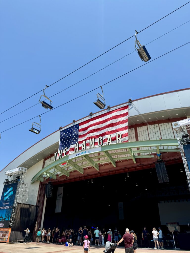 People gather outside The Hangar at the OC Fair in Costa Mesa, a large open building with an American flag hanging above its entrance. Overhead, several empty ski lift chairs glide across the clear blue sky.