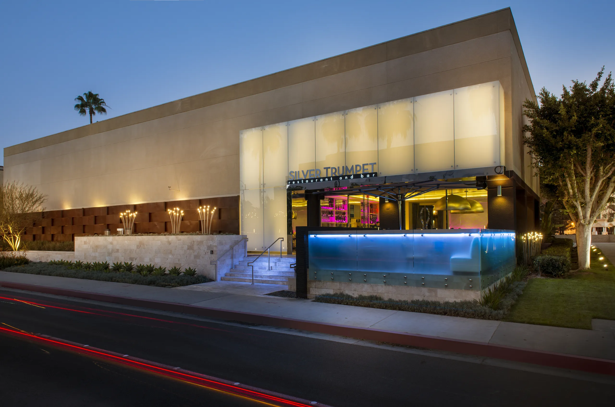 Modern building at dusk with the Silver Trumpet sign, illuminated steps, glass walls, and blue accent lighting. Palm trees and streaks of car lights are visible in the foreground and background, capturing the vibrant atmosphere surrounding Silver Trumpet Restaurant Costa Mesa.