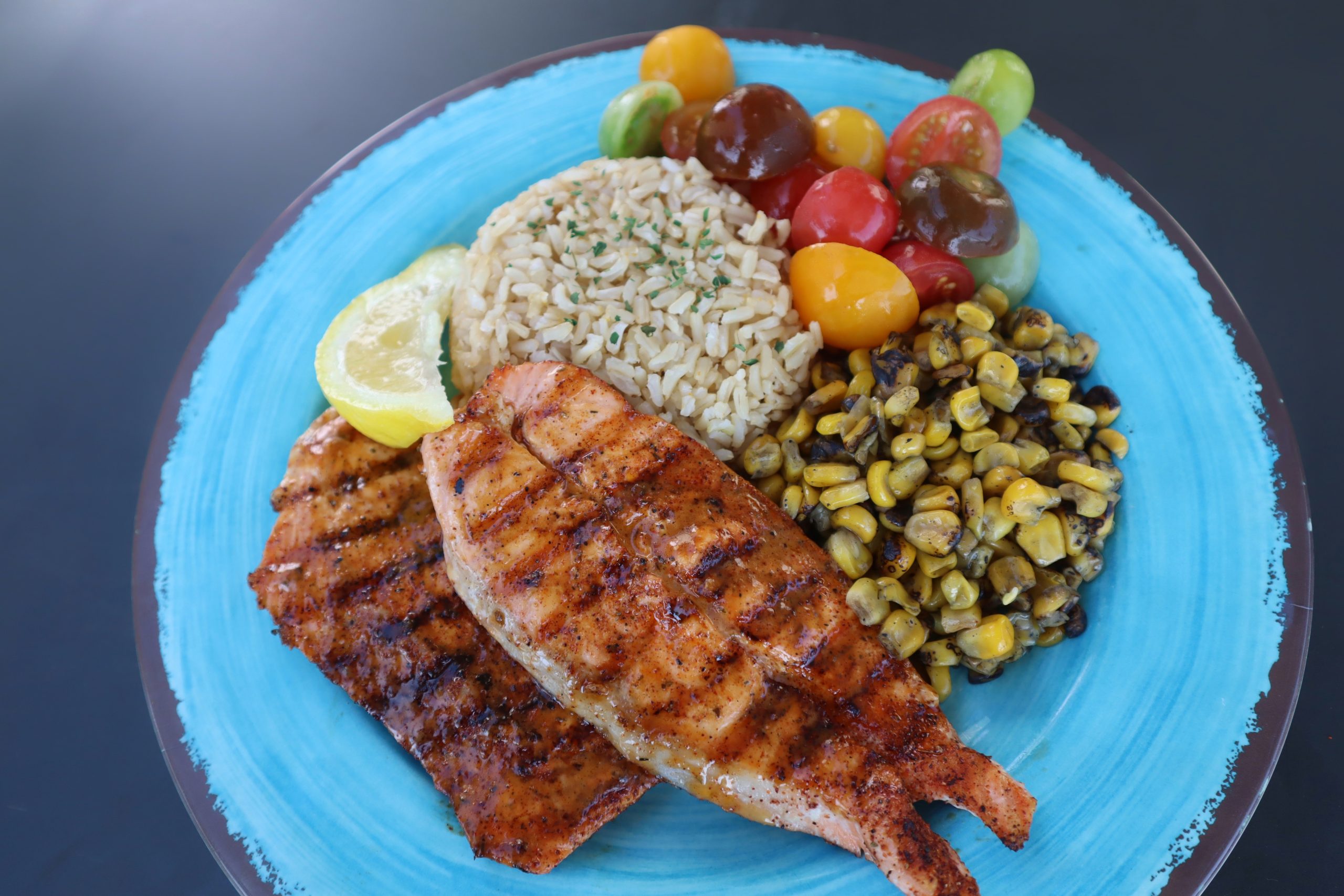 Two grilled fish fillets on a blue plate with brown rice, a lemon wedge, roasted corn, and colorful cherry tomatoes arranged beside each other.