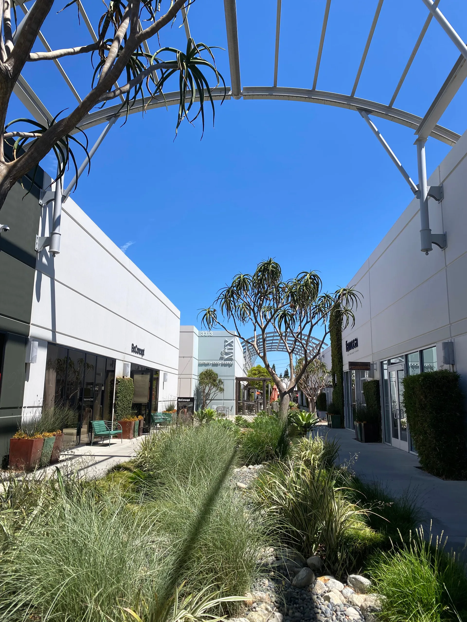 A modern outdoor shopping plaza with white buildings, glass storefronts, green landscaping, and tall trees under a clear blue sky. An arched metal canopy is overhead.