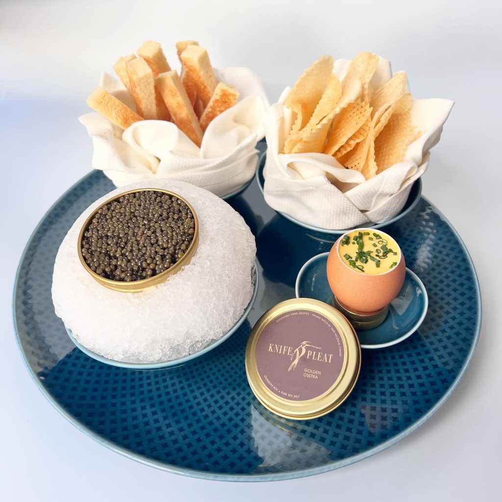 A blue plate holds a tin of caviar on ice, a soft-boiled egg topped with herbs, breadsticks, and thin crackers in white napkins. A closed tin labeled KNIFE & PLEAT is also on the plate.