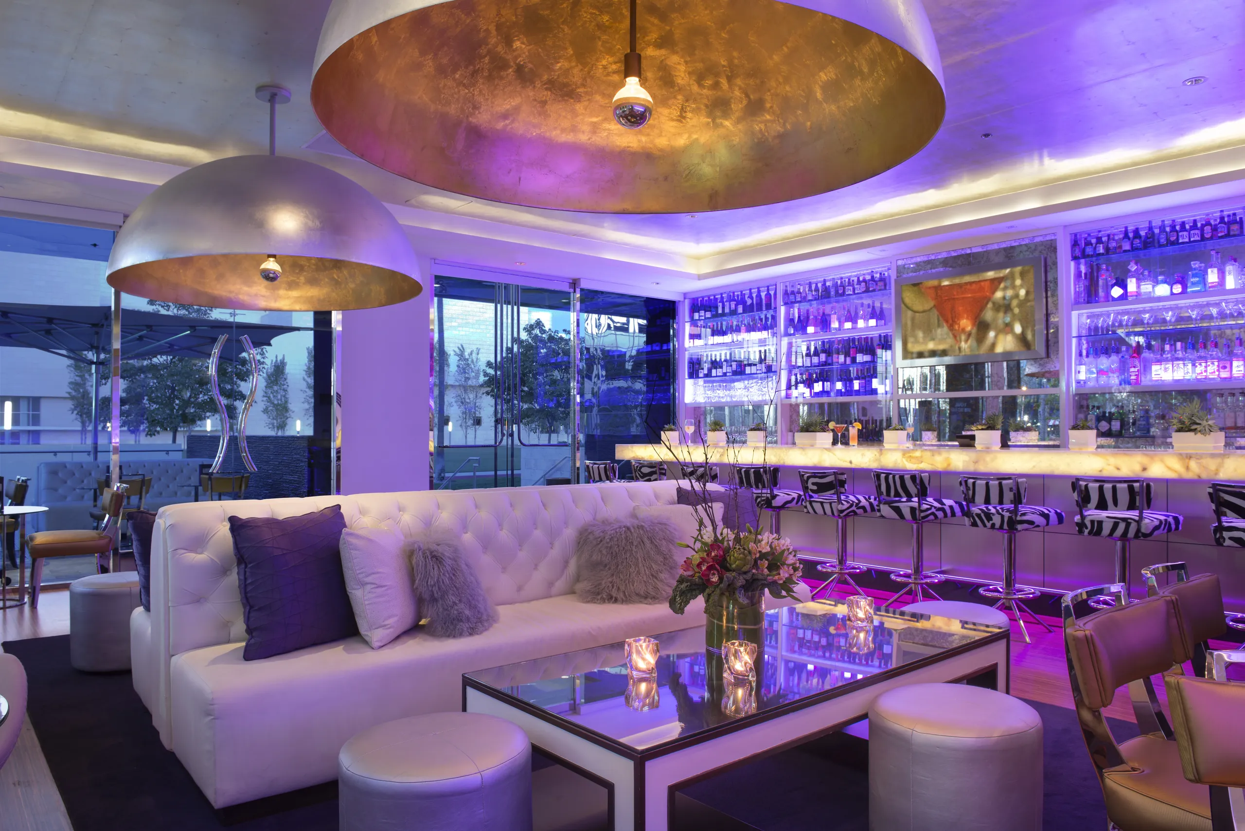 A modern lounge bar, reminiscent of the chic ambiance found at Silver Trumpet Restaurant Costa Mesa, features a white tufted sofa, glass tables, plush stools, large gold dome lights, and a backlit bar with bottles and zebra-patterned barstools. Purple lighting creates a stylish, upscale atmosphere.