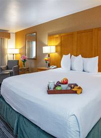 A neatly made hotel bed at Ramada Costa Mesa with white linens and three pillows, featuring a tray with fruit, a cup, and a napkin. The room offers warm lighting, a wooden headboard, a mirror, and a sitting area with flowers.
