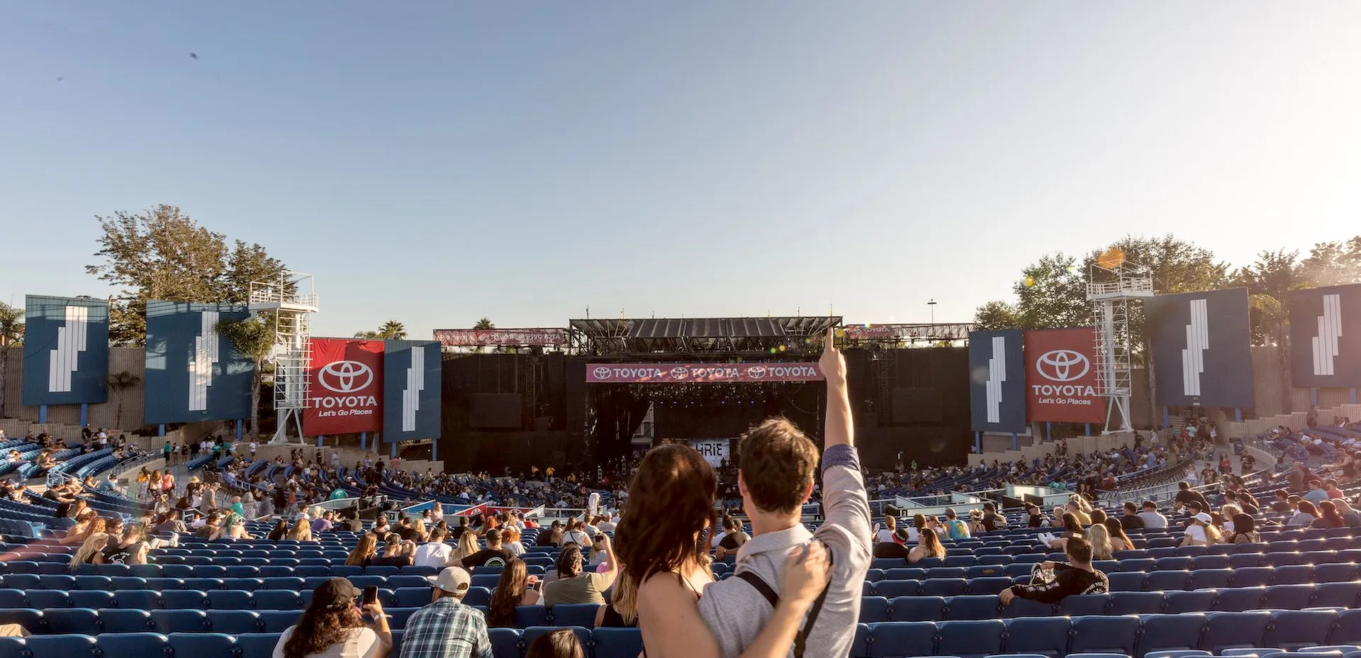 A couple stands embracing and pointing, facing an outdoor amphitheater with blue seats and a stage ahead. Large Toyota banners are displayed, and people are scattered throughout the audience on a sunny day—perfect for a Costa Mesa travel blog adventure.