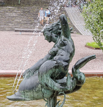 A bronze statue of a mermaid holding a fish, acting as a fountain with water spraying, is situated in a pond with stone steps and people in the background along the Costa Mesa Art Walk.