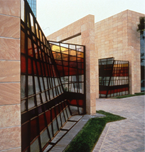 Modern building with tan stone walls and large, slanted windows featuring a grid of tinted glass panels in brown and red, set along a paved walkway with grass patches—perfectly capturing the spirit of the Costa Mesa Art Walk.