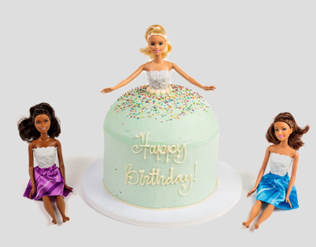 Three dolls are posed around a round birthday cake with light green frosting, colorful sprinkles, and “Happy Birthday!” written on it. The doll in the center appears to be part of the cake design.