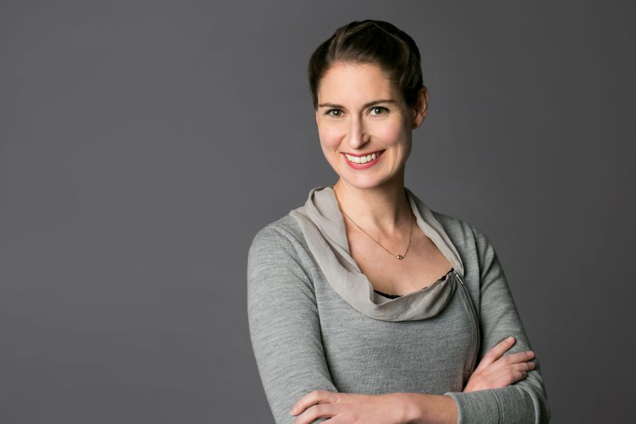 A woman with brown hair pulled back, wearing a grey long-sleeve top and a necklace, smiles with her arms crossed in front of a plain grey background.