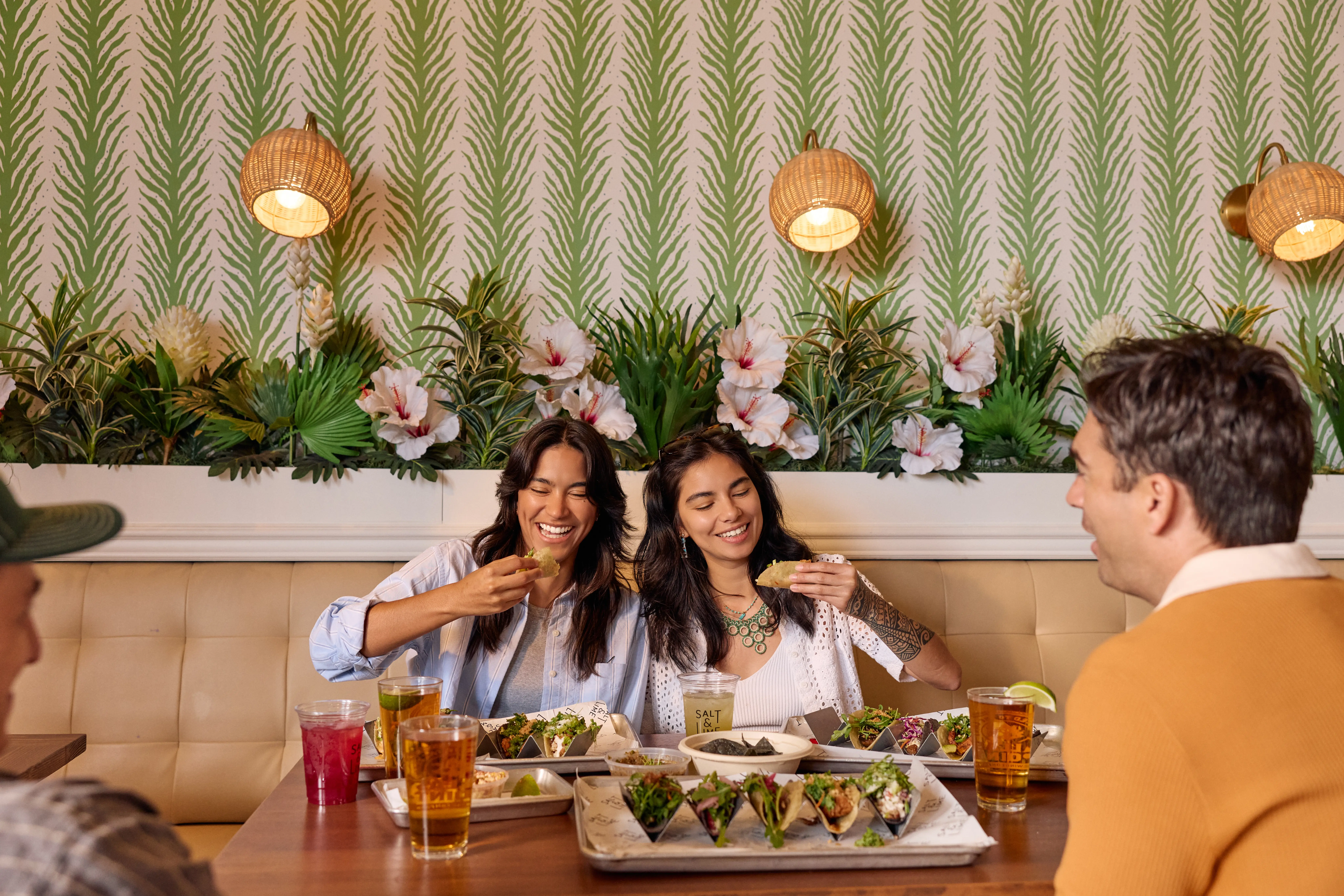Four people sit around a table in a tropical-themed restaurant, laughing and enjoying drinks and food, including tacos and guacamole. Two women in the center are smiling while eating, with decorative greenery and flowers on the wall behind them.