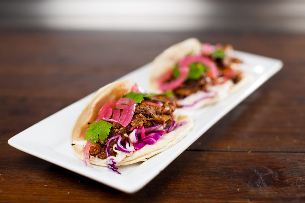 Two soft tacos filled with shredded meat, purple cabbage, pickled red onions, and cilantro, served on a rectangular white plate on a wooden table.