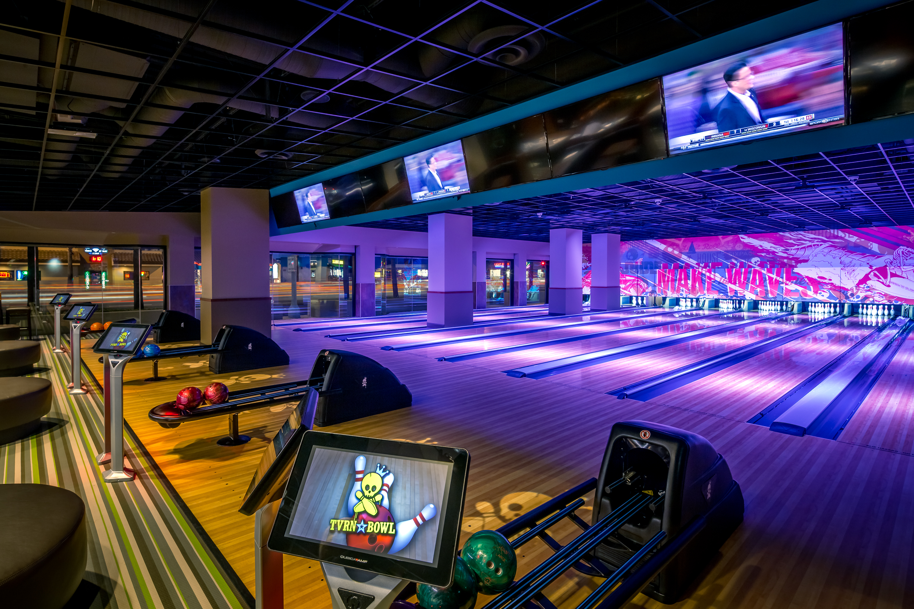A modern bowling alley at Triangle Square Costa Mesa with several empty lanes, colorful bowling balls, digital score screens, and TVs displaying sports above the lanes. The space is brightly lit with vivid, colorful decor.