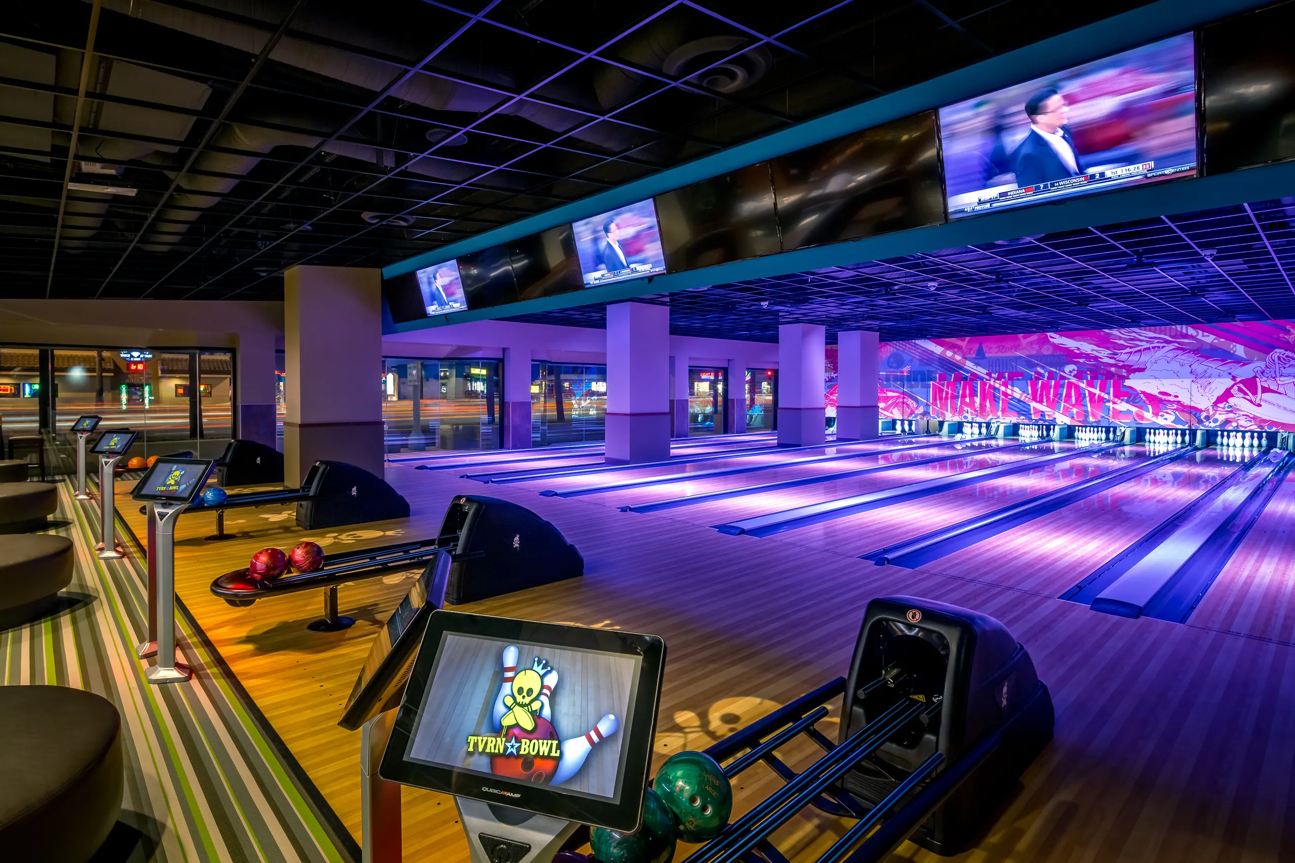 A modern bowling alley at Triangle Square Costa Mesa with several empty lanes, colorful bowling balls, digital score screens, and TVs displaying sports above the lanes. The space is brightly lit with vivid, colorful decor.