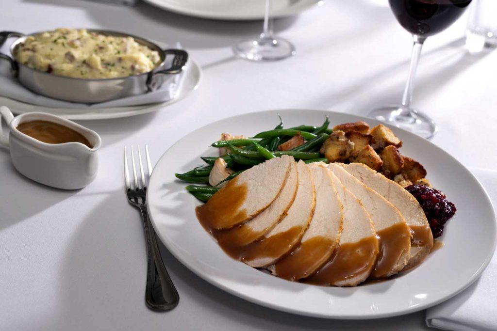 A plate with sliced turkey topped with brown gravy, green beans, stuffing, and cranberry sauce. Nearby are a fork, a dish of mashed potatoes, a small gravy boat, and a glass of red wine on a white tablecloth.