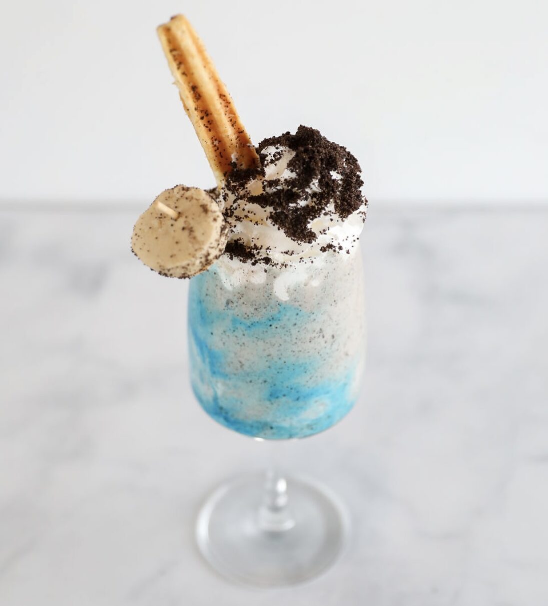 A tall glass filled with a blue and white swirled milkshake, topped with whipped cream, crushed cookies, a round cookie, and two churros, served on a marble surface at Silver Trumpet Restaurant Costa Mesa.