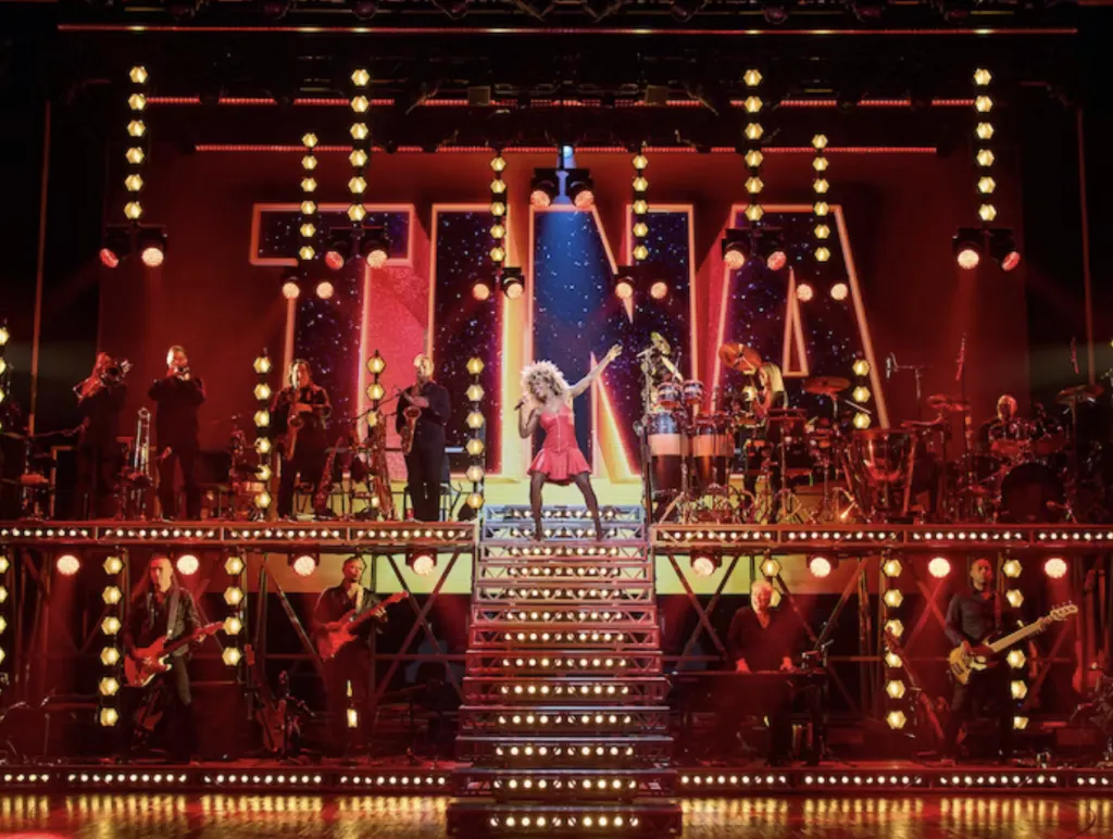 A performer in a red dress stands at the top of illuminated stairs, striking a dynamic pose on stage, with musicians playing instruments on both sides and large “TINA” letters lit up in the background.