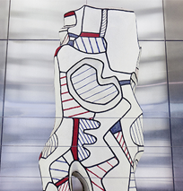 A white abstract sculpture with black, red, and blue outlined shapes and patterns stands in front of a metallic, tiled wall along the Costa Mesa Art Walk. The piece features dynamic curved and angular segments.