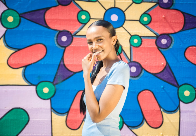A woman in a light blue dress smiles while standing in front of a colorful geometric mural, capturing the vibrant essence of solo travel in Costa Mesa with its red, blue, green, yellow, and purple circular and petal shapes.