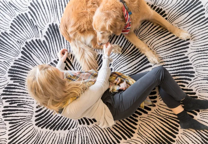 A woman with blonde hair sits on a patterned black-and-white rug, leaning toward a golden retriever. The dog lies beside her, wearing a patterned scarf around its neck.