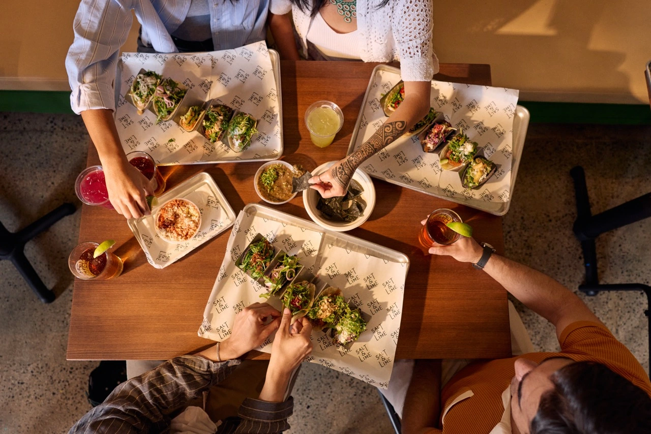 Four people sit around a wooden table, sharing tacos, guacamole, salsa, and drinks. The photo is taken from above, showing hands reaching for food on paper-lined trays. The setting appears casual and lively.