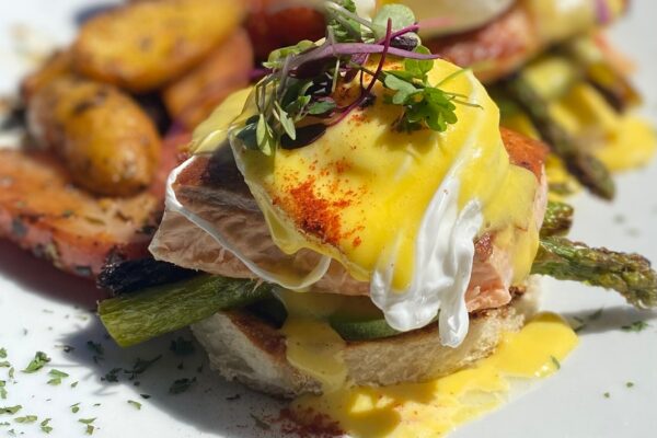 At Tres Muchachos Costa Mesa, enjoy Eggs Benedict on toasted bread with salmon, asparagus, poached eggs, and hollandaise sauce, topped with microgreens. Roasted potatoes and tomatoes complete the plate for a satisfying brunch.