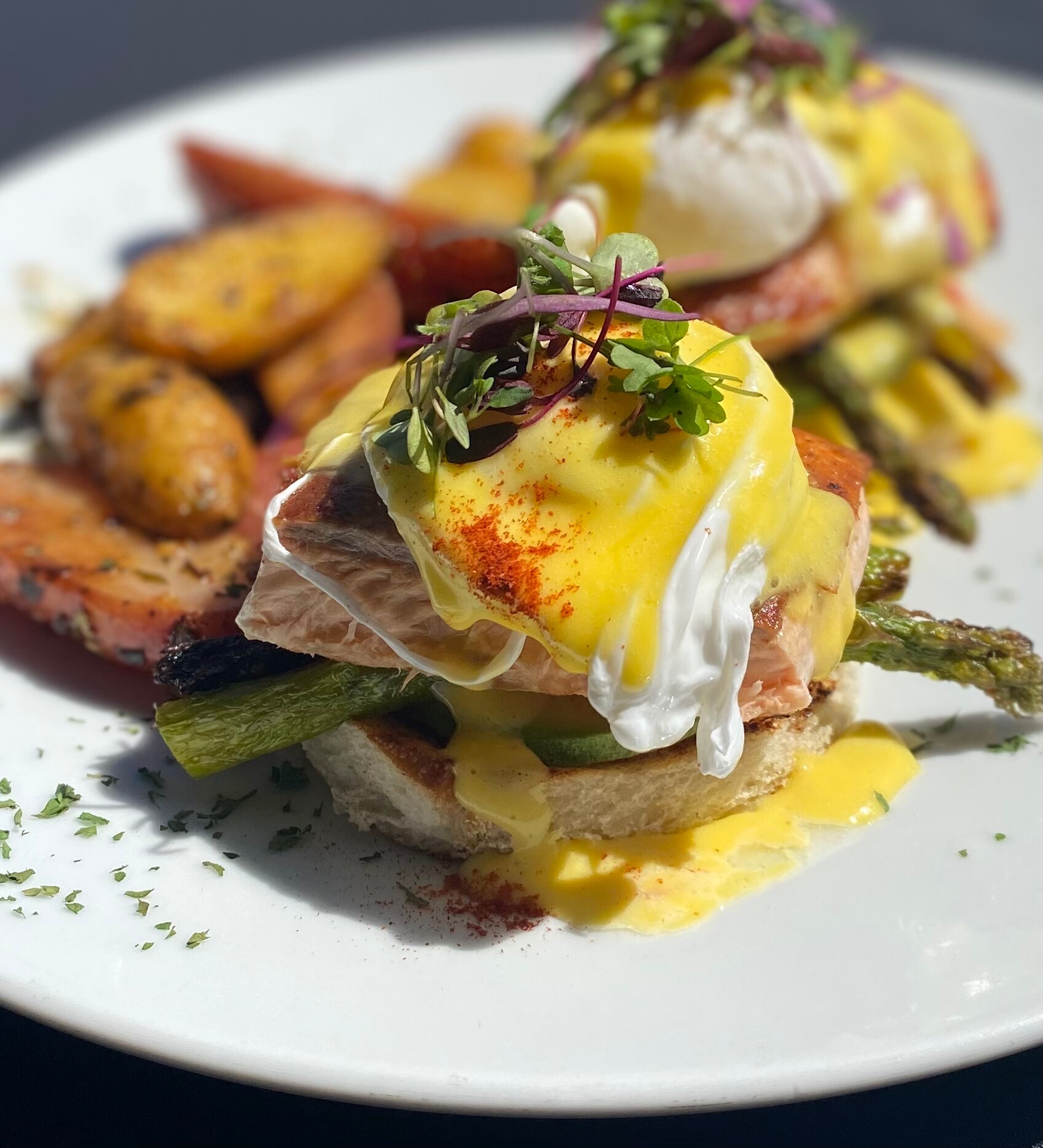 At Tres Muchachos Costa Mesa, enjoy Eggs Benedict on toasted bread with salmon, asparagus, poached eggs, and hollandaise sauce, topped with microgreens. Roasted potatoes and tomatoes complete the plate for a satisfying brunch.