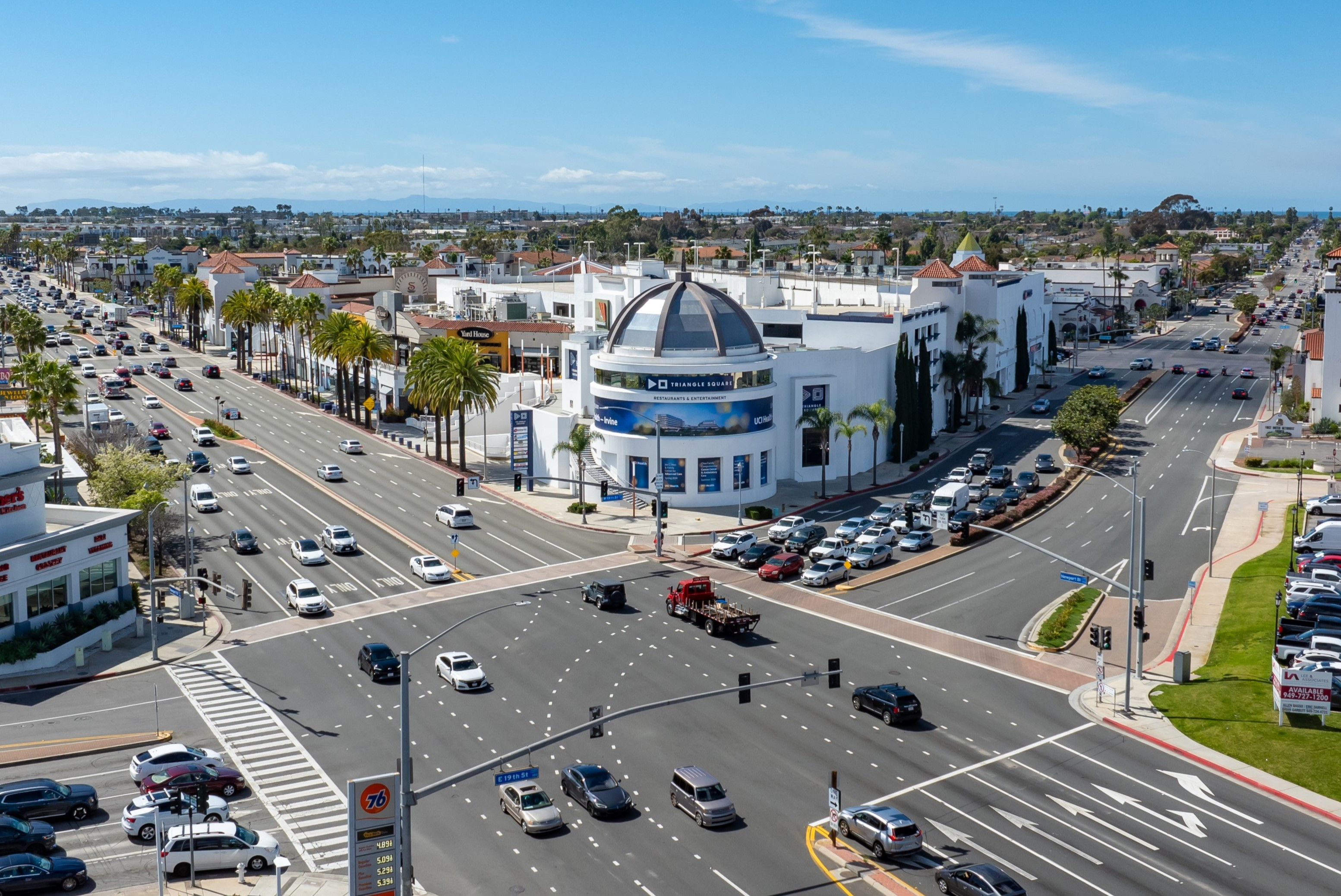 A busy intersection in Costa Mesa’s urban area with palm trees, many cars, and large white buildings—like those near Triangle Square—under a blue sky. Traffic signals and crosswalks are visible.