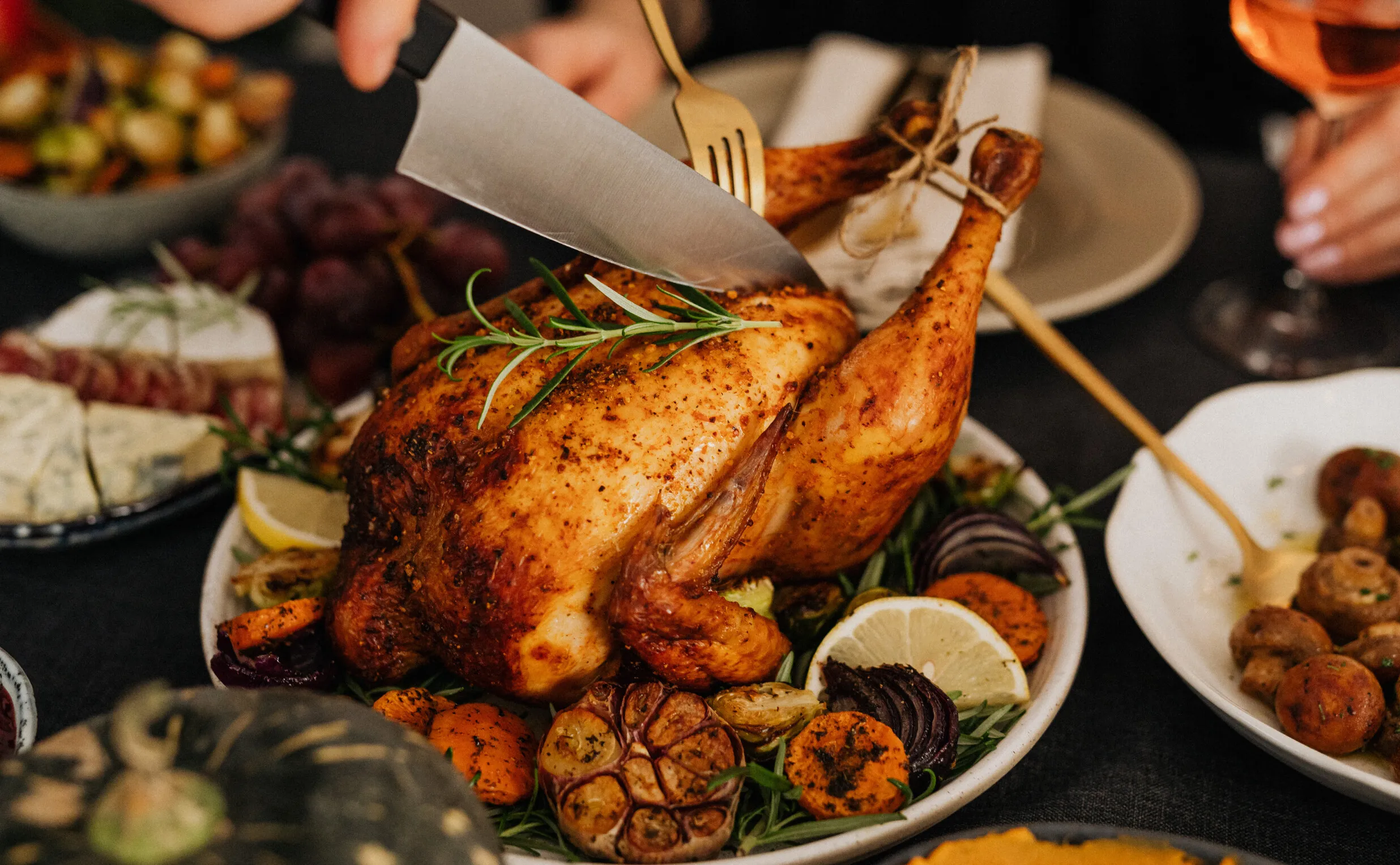 A person carves a roasted chicken garnished with rosemary, surrounded by lemon slices, roasted garlic, and vegetables—an inviting scene perfect for a costa mesa travel blog capturing festive meals and local flavors with wine in the background.