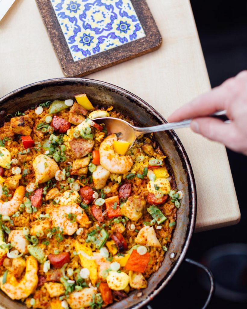 A hand scoops a spoonful of colorful paella from a pan, filled with shrimp, sausage, chicken, peppers, and rice. A decorative tile sits on a cork mat nearby.