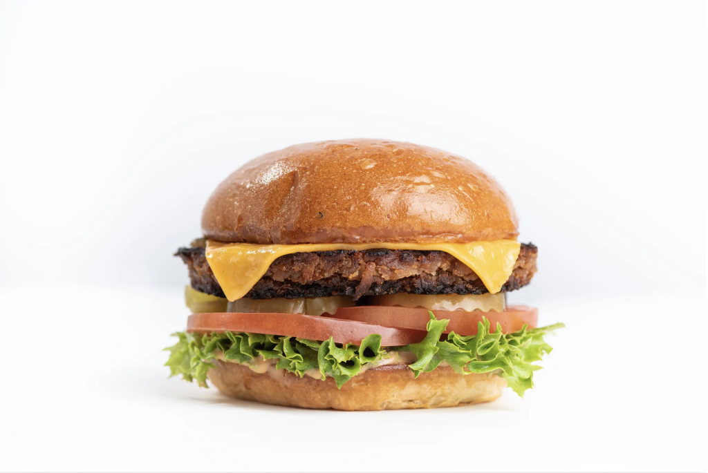 A cheeseburger with a golden bun, a slice of cheddar cheese, lettuce, tomato, pickles, and a crispy patty, all stacked neatly on a white background.