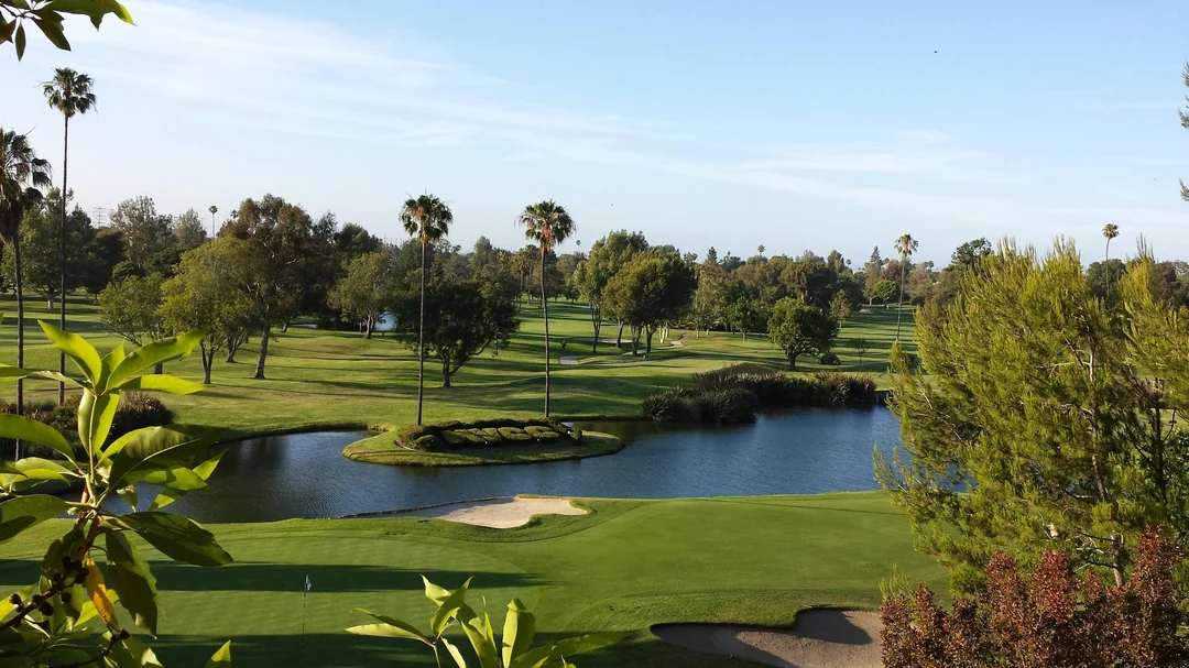 A green golf course with sand bunkers and a small pond, surrounded by trees and palm trees under a clear blue sky.