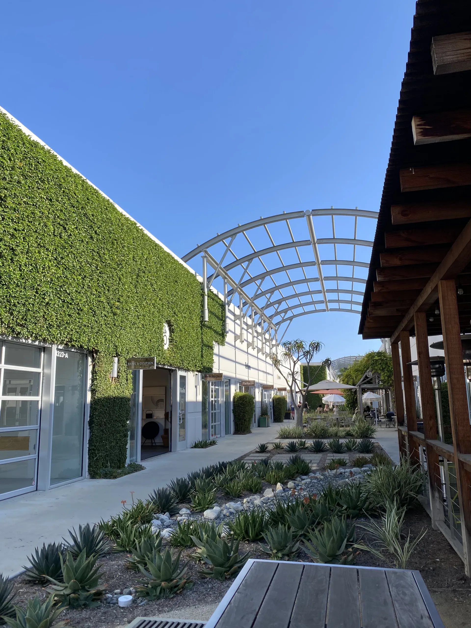 A modern outdoor walkway at SOCO OC Mix Costa Mesa features a curved metal canopy, lush ivy covering one building, and a landscaped garden with succulents and rocks under a clear blue sky.