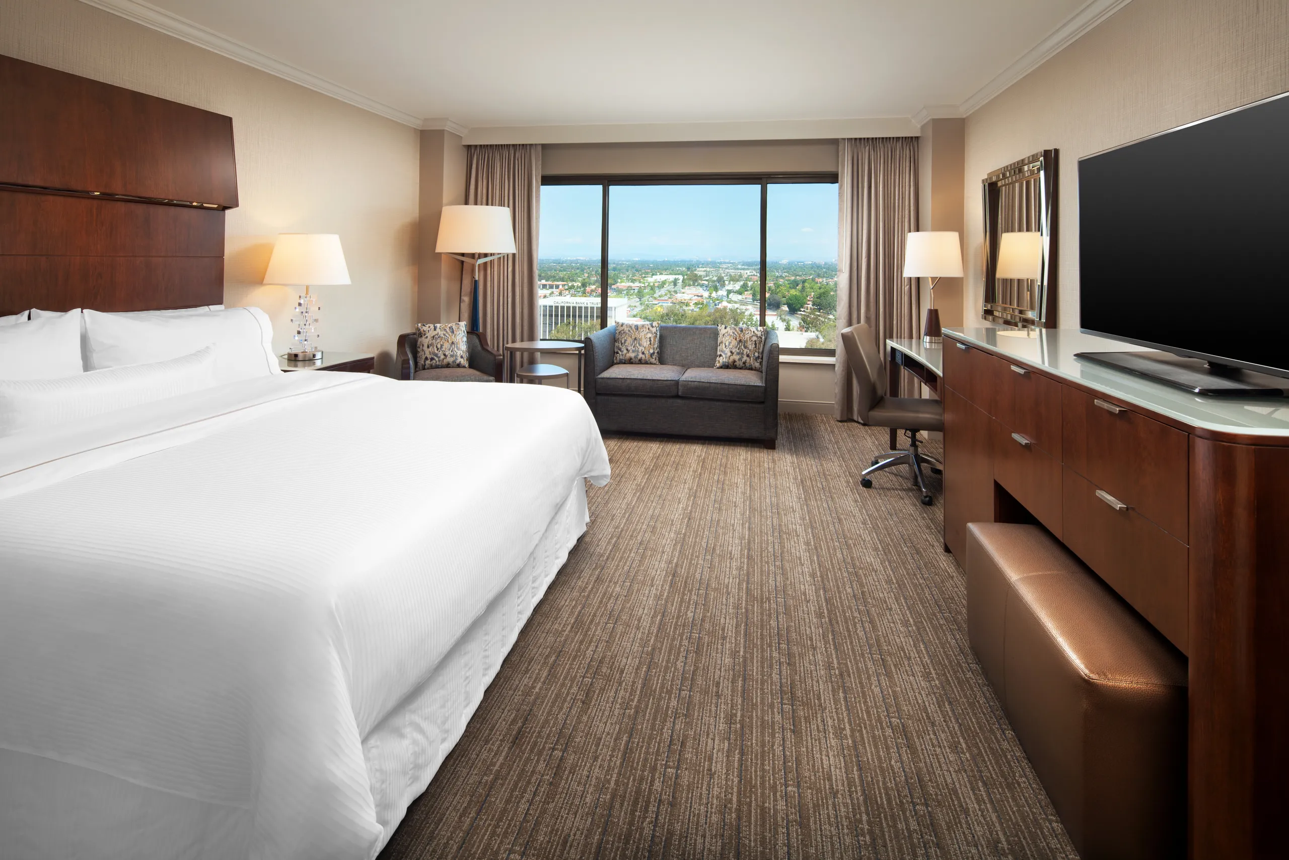 Spacious Westin South Coast Plaza hotel room with a large bed, sofa, desk with chair, flat-screen TV, and large window with city view; neutral tones and contemporary furniture.