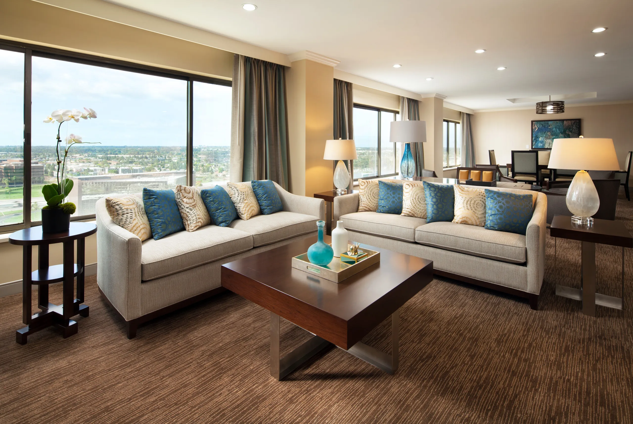 A modern living room at the Westin South Coast Plaza features large windows, two beige sofas with blue and beige pillows, a wooden coffee table, lamps, an orchid plant, and a dining area in the background bathed in natural light.