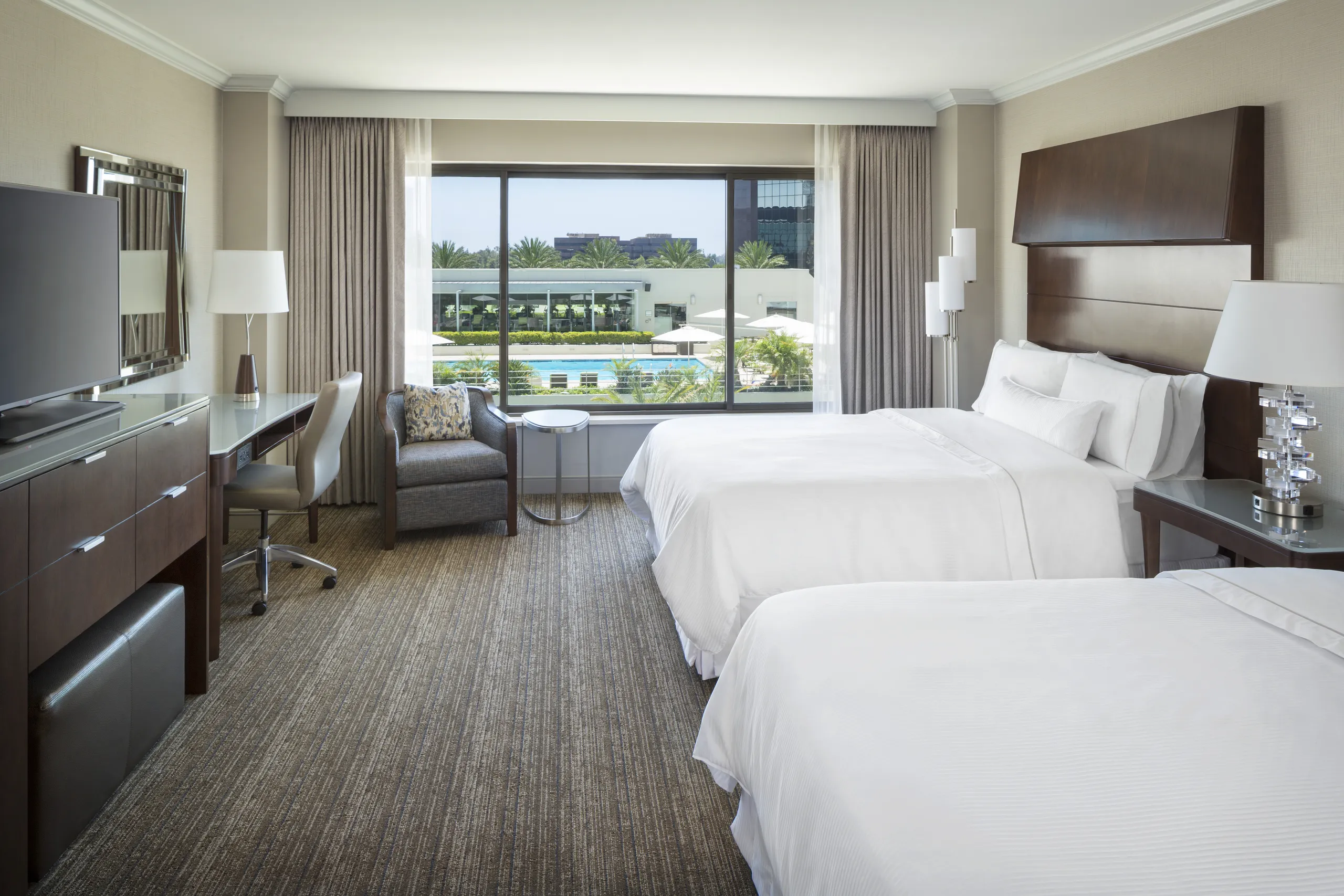 A modern Westin South Coast Plaza hotel room with two double beds, a large window overlooking the pool and trees, a desk and chair, armchair, TV on a cabinet, and bedside lamps. The room is bright and tidy.