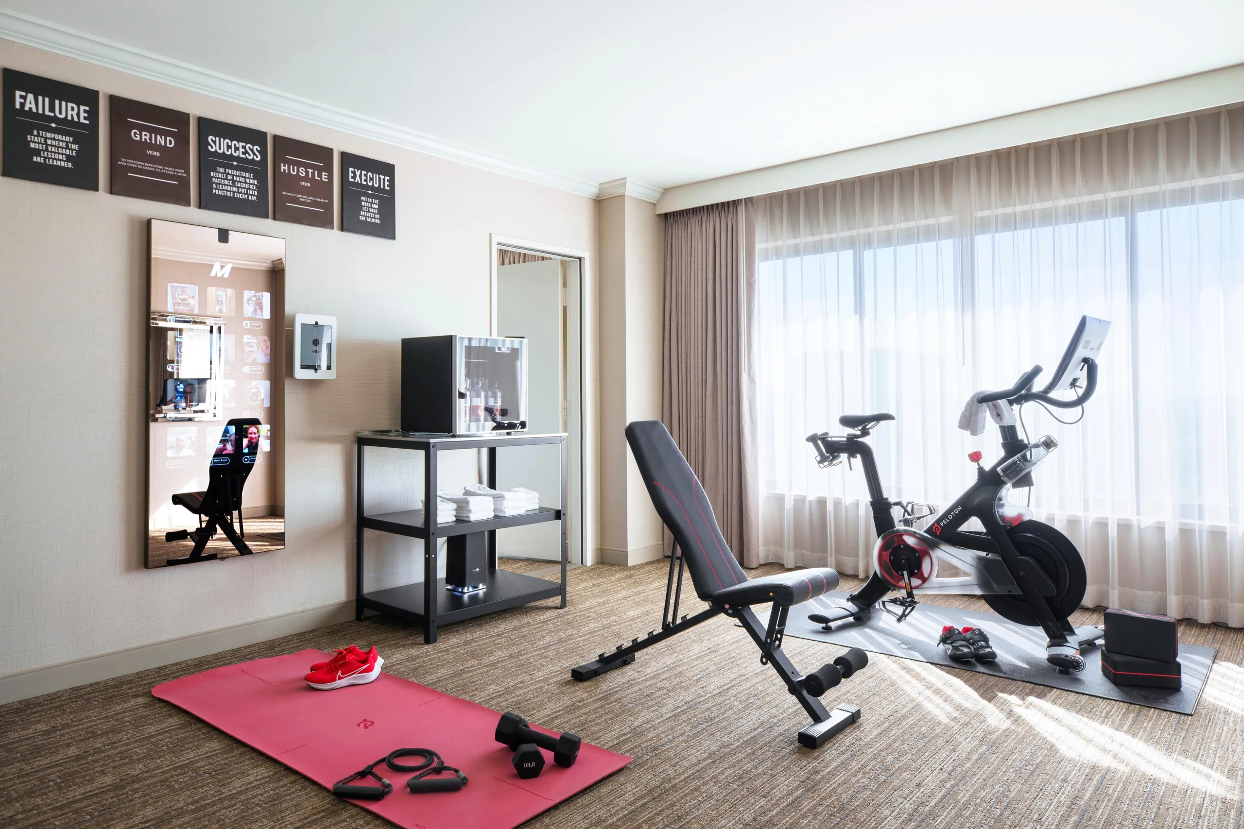 A bright home gym room at Westin South Coast Plaza features an exercise bike, weight bench, red yoga mat, dumbbells, resistance bands, towels, a mirror, and motivational signs beside a large window with sheer curtains.