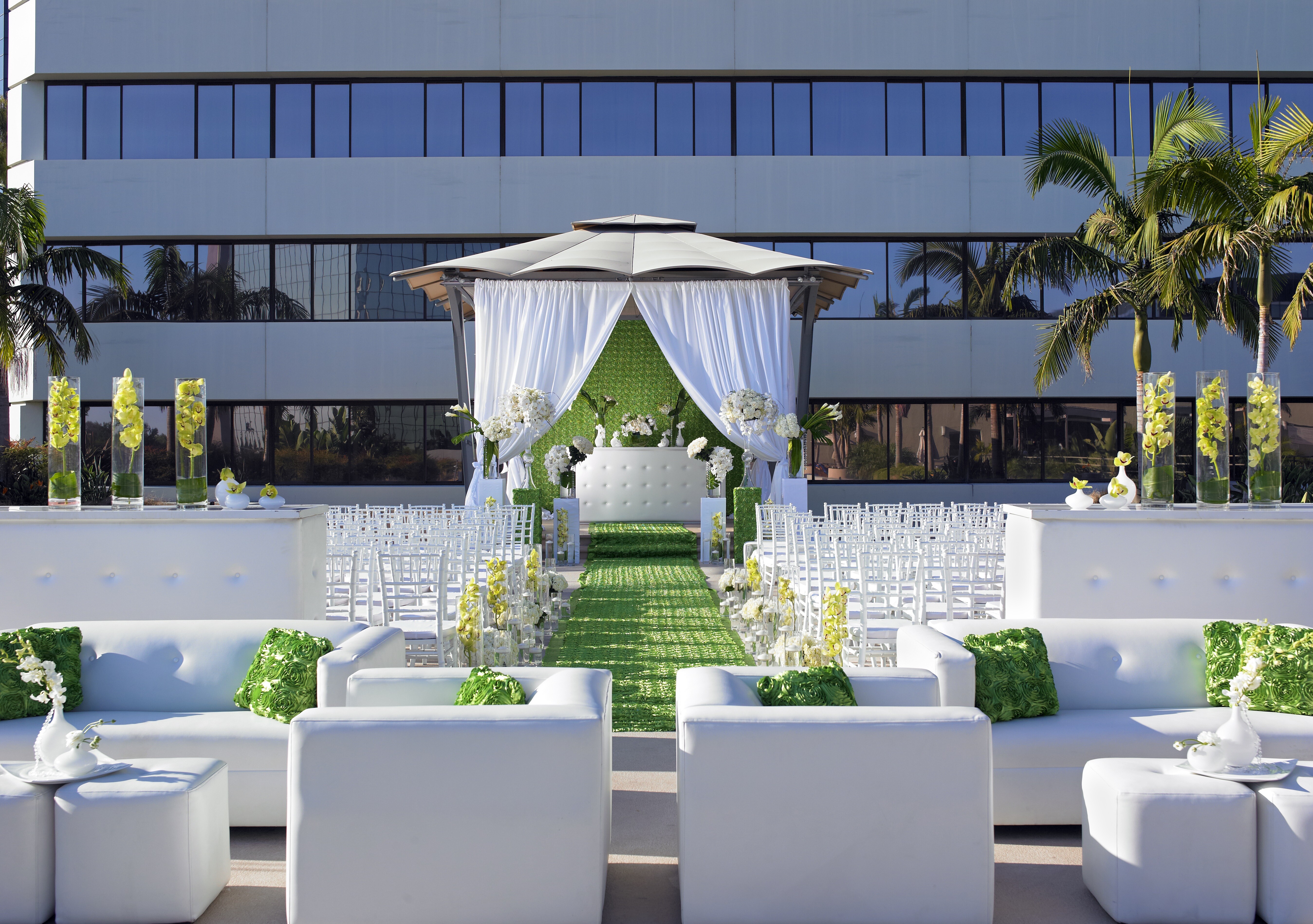 An outdoor wedding setup at Westin South Coast Plaza features white sofas, chairs, and green accents, including a green aisle runner and pillows. A gazebo with white drapes stands at the end, framed by palm trees and a modern glass building.