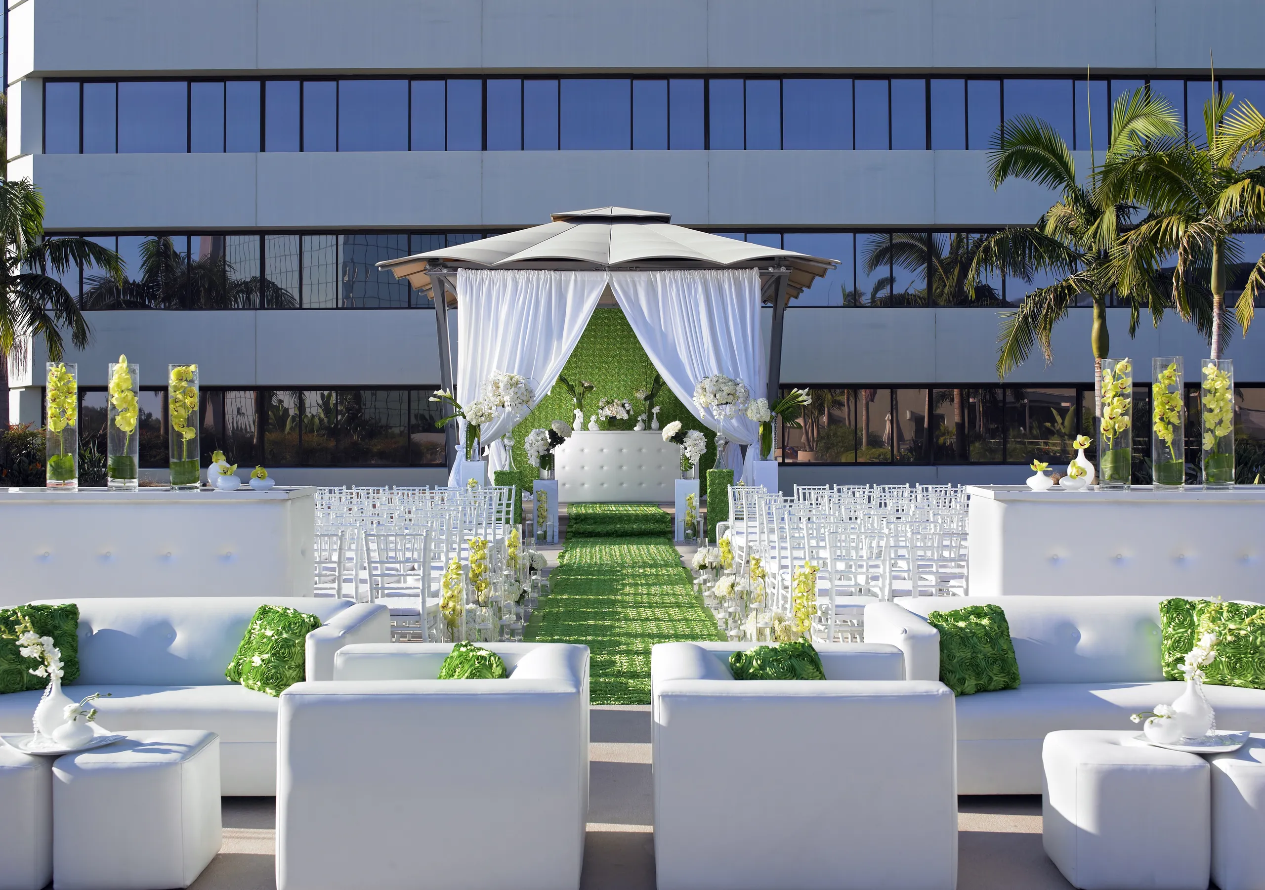 An outdoor wedding setup at Westin South Coast Plaza features white sofas, chairs, and green accents, including a green aisle runner and pillows. A gazebo with white drapes stands at the end, framed by palm trees and a modern glass building.