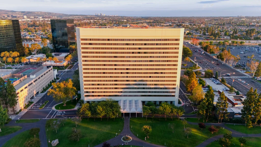 Aerial view of the Westin South Coast Plaza, a tall, rectangular office building surrounded by trees, roads, and parked cars in a cityscape during the late afternoon.