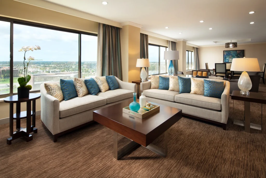 A spacious, modern living room at Westin South Coast Plaza features large windows, beige sofas with blue and cream pillows, a wooden coffee table, lamps, and cityscape views. A dining area is visible in the background.