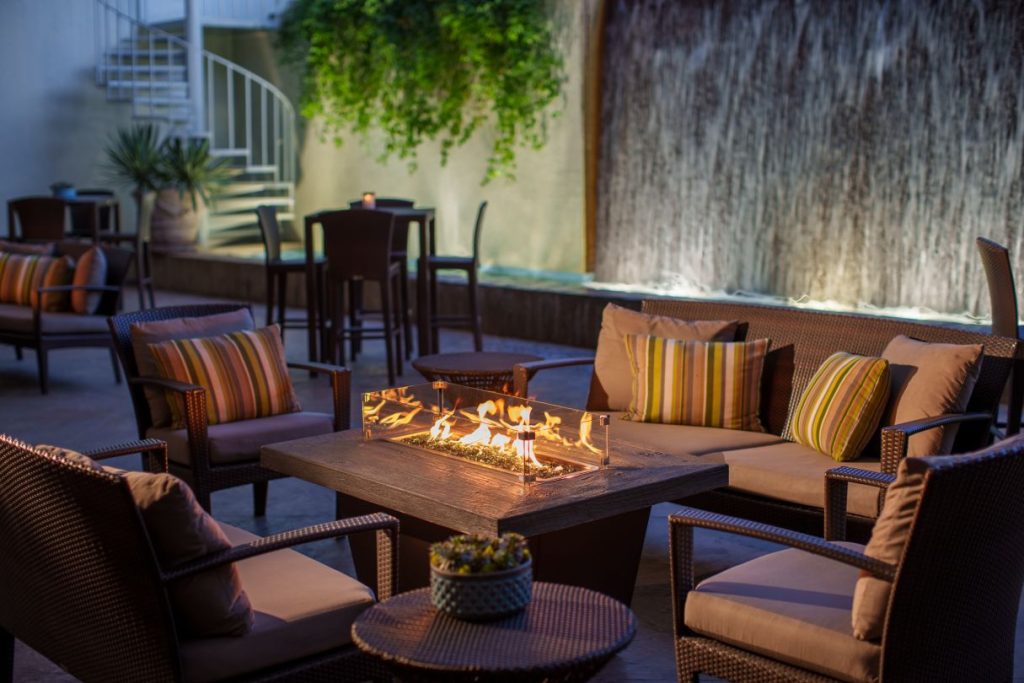 An outdoor patio features wicker chairs with colorful striped cushions arranged around a modern fire pit. In the background, a vertical water feature and lush greenery create a relaxing atmosphere.