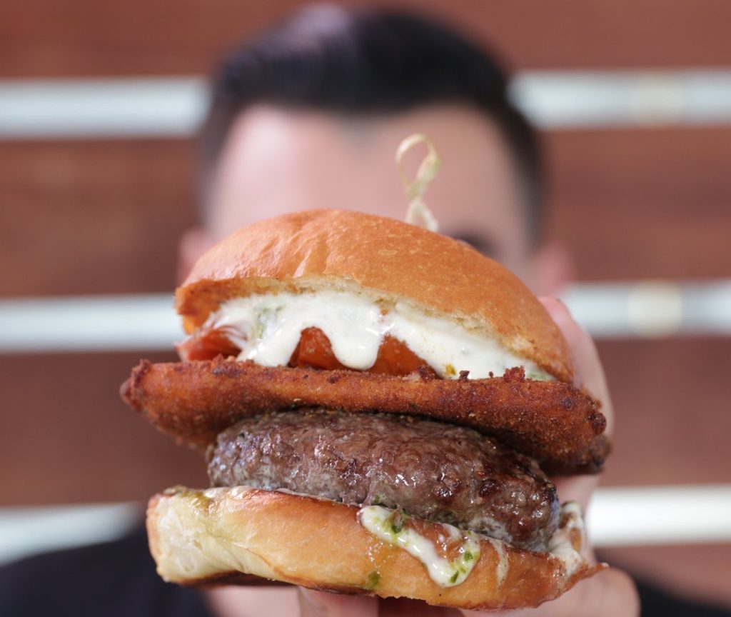 A person holds up a close-up of a hamburger with a beef patty, fried topping, melted cheese, sauce, and a brioche bun, with the background slightly blurred.