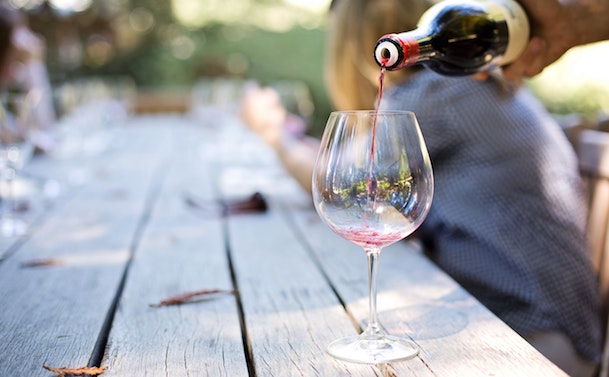 A person pours red wine from a bottle into a glass on a weathered wooden outdoor table, with more glasses and blurred figures in the background.