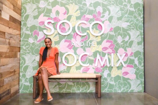 A woman in an orange dress sits smiling on a wooden bench in front of a floral wall with large white letters reading “SOCO AND THE OC MIX.”.
