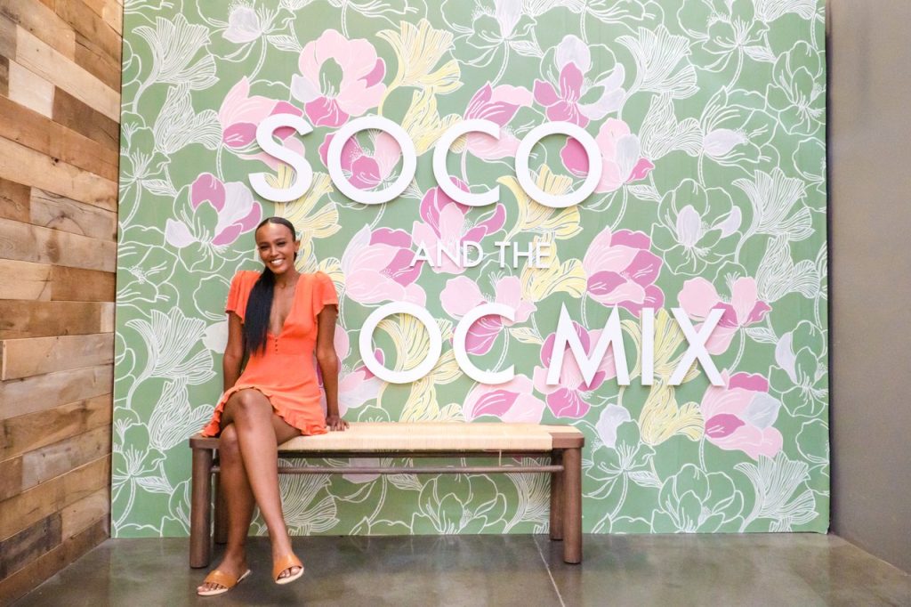 A woman in an orange dress sits smiling on a wooden bench in front of a floral wall with large white letters reading “SOCO AND THE OC MIX.”.