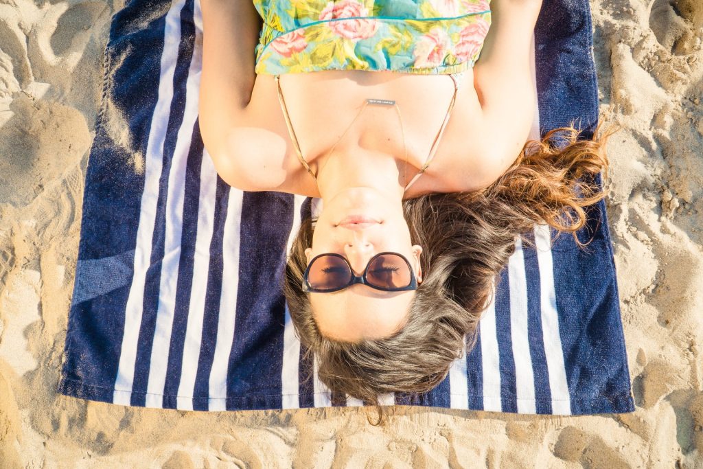 A woman wearing sunglasses and a floral dress lies on a blue and white striped towel on sandy beach, relaxing in the sun with her hair spread out.