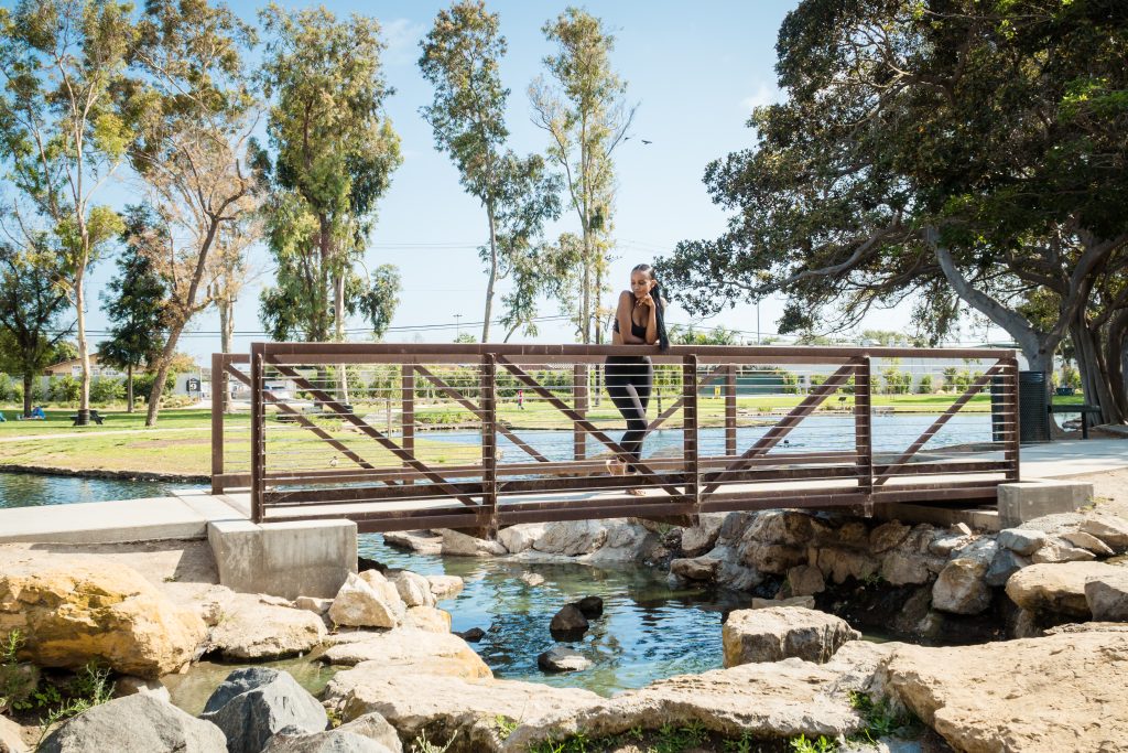 A woman stands on a wooden footbridge over a small stream in TeWinkle Park, Costa Mesa, surrounded by trees and rocks on a sunny day.
